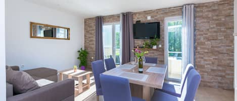 A modern living room with a sofa, a dining table with chairs, a TV mounted on the wall, and a balcony with a view.