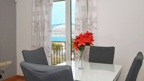 Un coin repas avec une table et des chaises, un vase rempli de fleurs rouges et une vue sur la mer par une fenêtre ornée de rideaux.