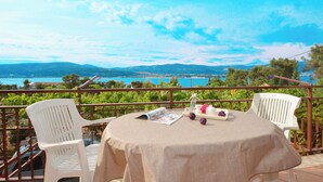 Une table ronde recouverte d’une nappe beige, deux chaises blanches et une vue sur un paysage côtier avec des montagnes et une étendue d’eau.