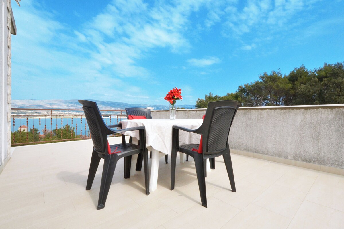 A terrace with a table and chairs, a vase with red flowers, and a view of the sea and mountains.