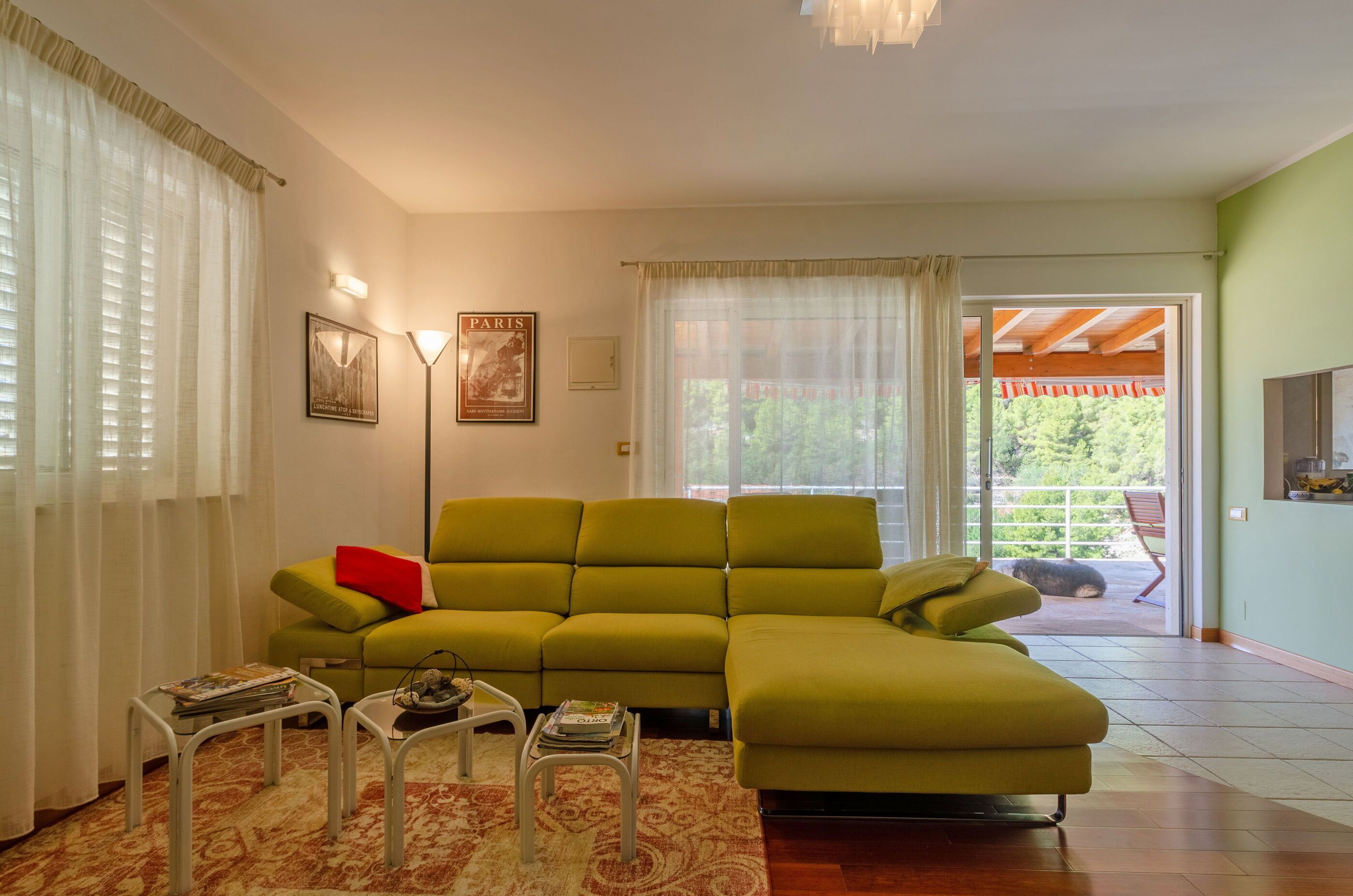 A living room with a green sofa, a small table with magazines, a lamp, and a view of a balcony with a red chair.
