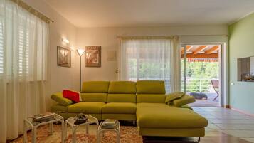 A living room with a green sofa, a small table with magazines, a lamp, and a view of a balcony with a red chair.