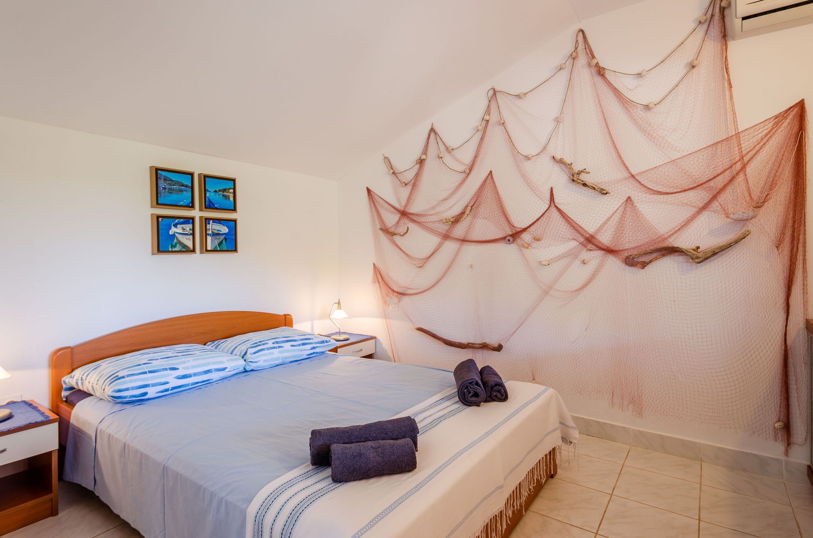 A neatly made bed with a blue and white striped pillow, a pair of rolled towels, and a decorative net hanging on the wall.