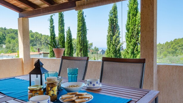 Une table en bois surmontée d’une nappe bleue, une tasse blanche ornée de motifs floraux bleus, un verre, une lanterne, un bol de noix et une assiette de pain, le tout placé sur un balcon offrant une vue sur de grands arbres et un paysage lointain.