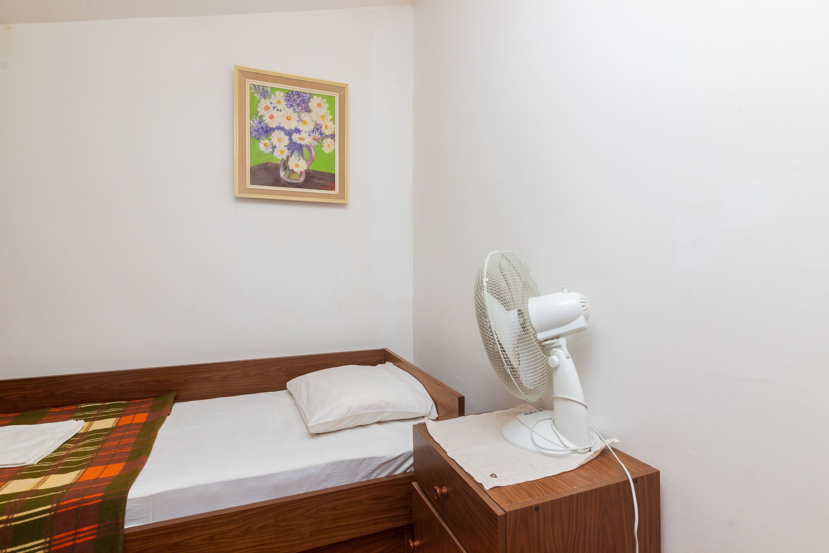 Ein Einzelbett mit Holzbettrahmen, einem weißen Kopfkissen, einer weißen Decke und einem kleinen Nachttischchen mit einem weißen Ventilator.