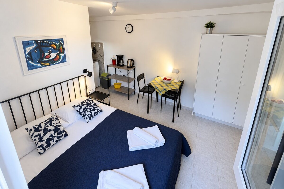 A compact room with a bed, a small dining table with chairs, a kitchenette, and a wardrobe.