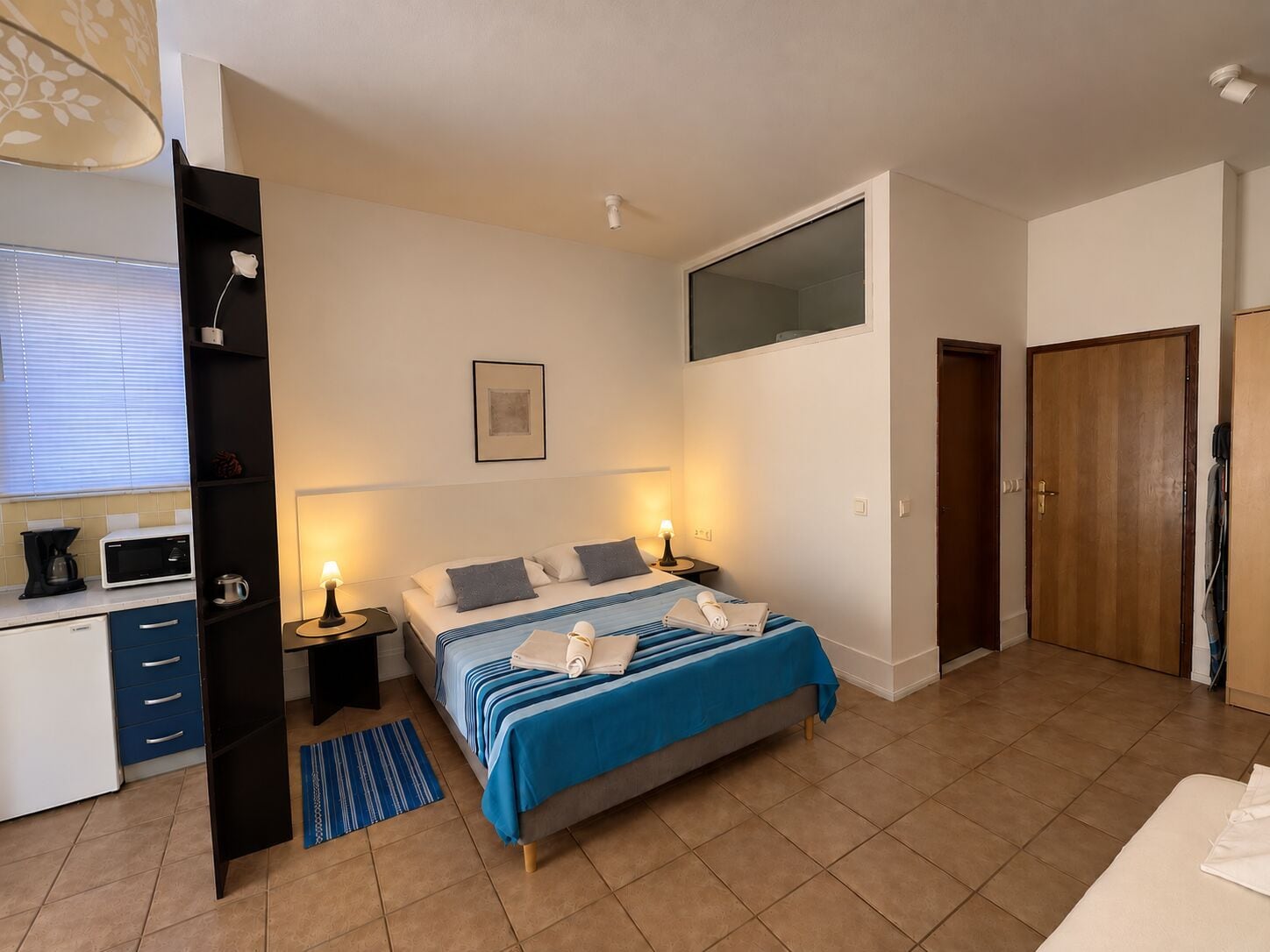 A bedroom with a bed, bedside tables, a desk with a computer, a window with blinds, and a door.