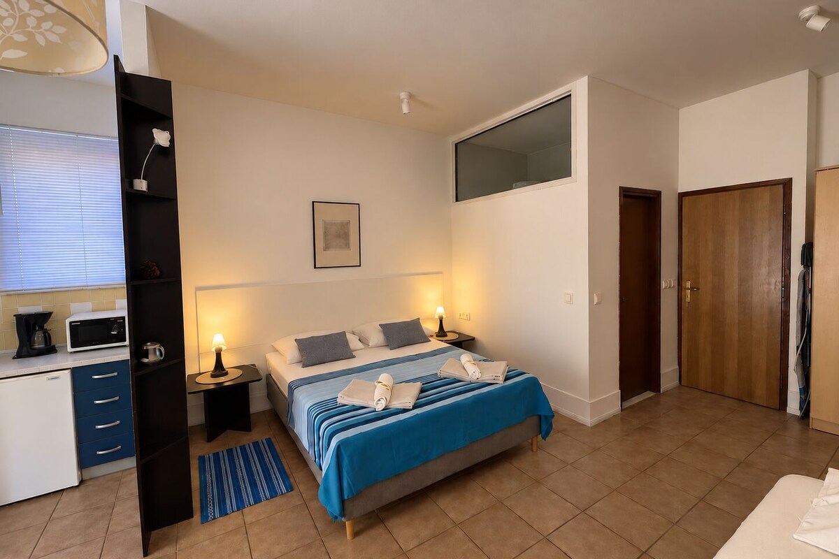 A bedroom with a bed, bedside tables, a desk with a computer, a window with blinds, and a door.