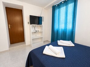 A hotel room with a bed, a TV on a stand, a door, and a window with curtains.