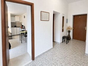 A hallway with a kitchen area visible through an open door, a framed notice on the wall, and a door with a vase of dried flowers. - Apartments Jugo i Bura (Omis)