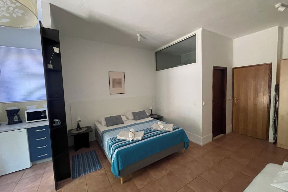 A bedroom with a bed, a bedside table, a nightstand, a window, a door, and a kitchenette area.