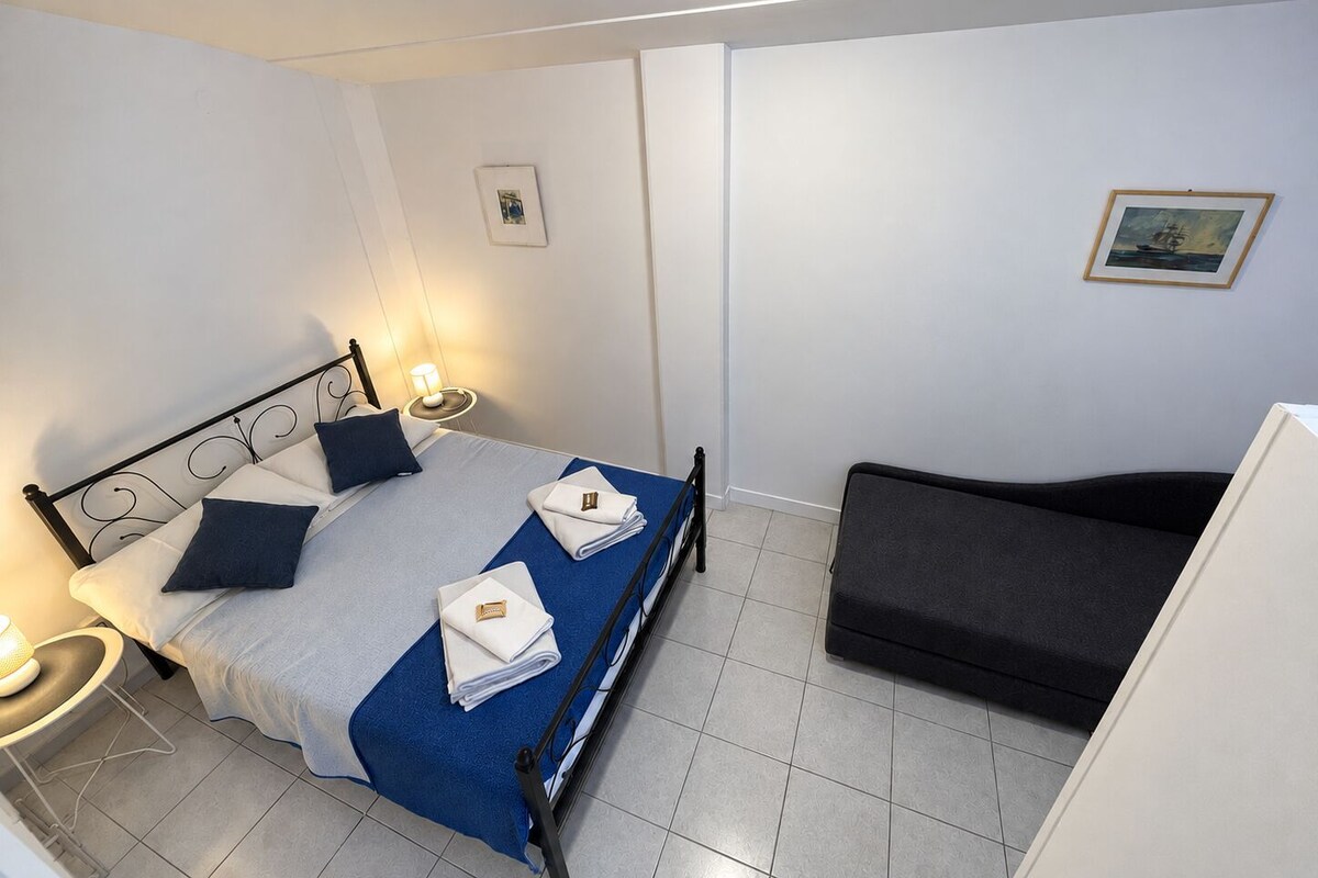 A single bed with a blue bedspread, two pillows, a bedside lamp, a small table, a dark sofa, and a framed picture on the wall.