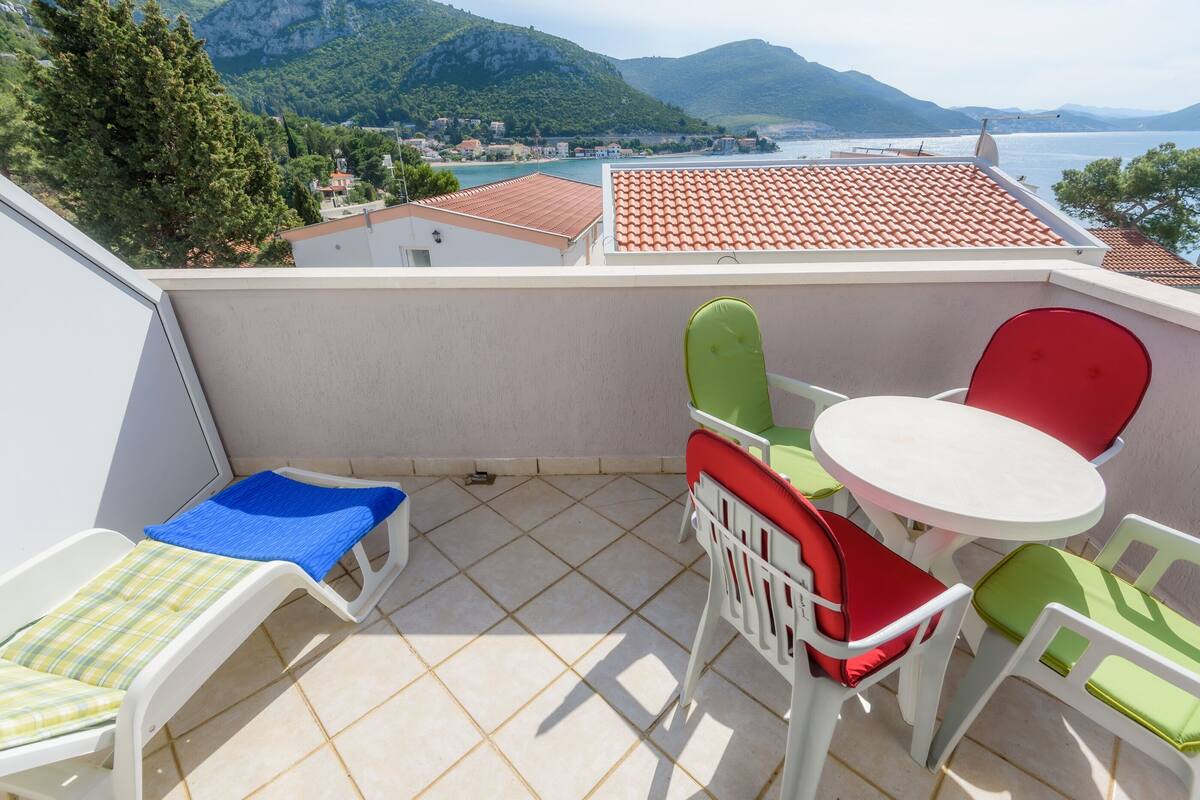 A balcony with a table and chairs, a small bed, and a view of a coastal town and mountains.