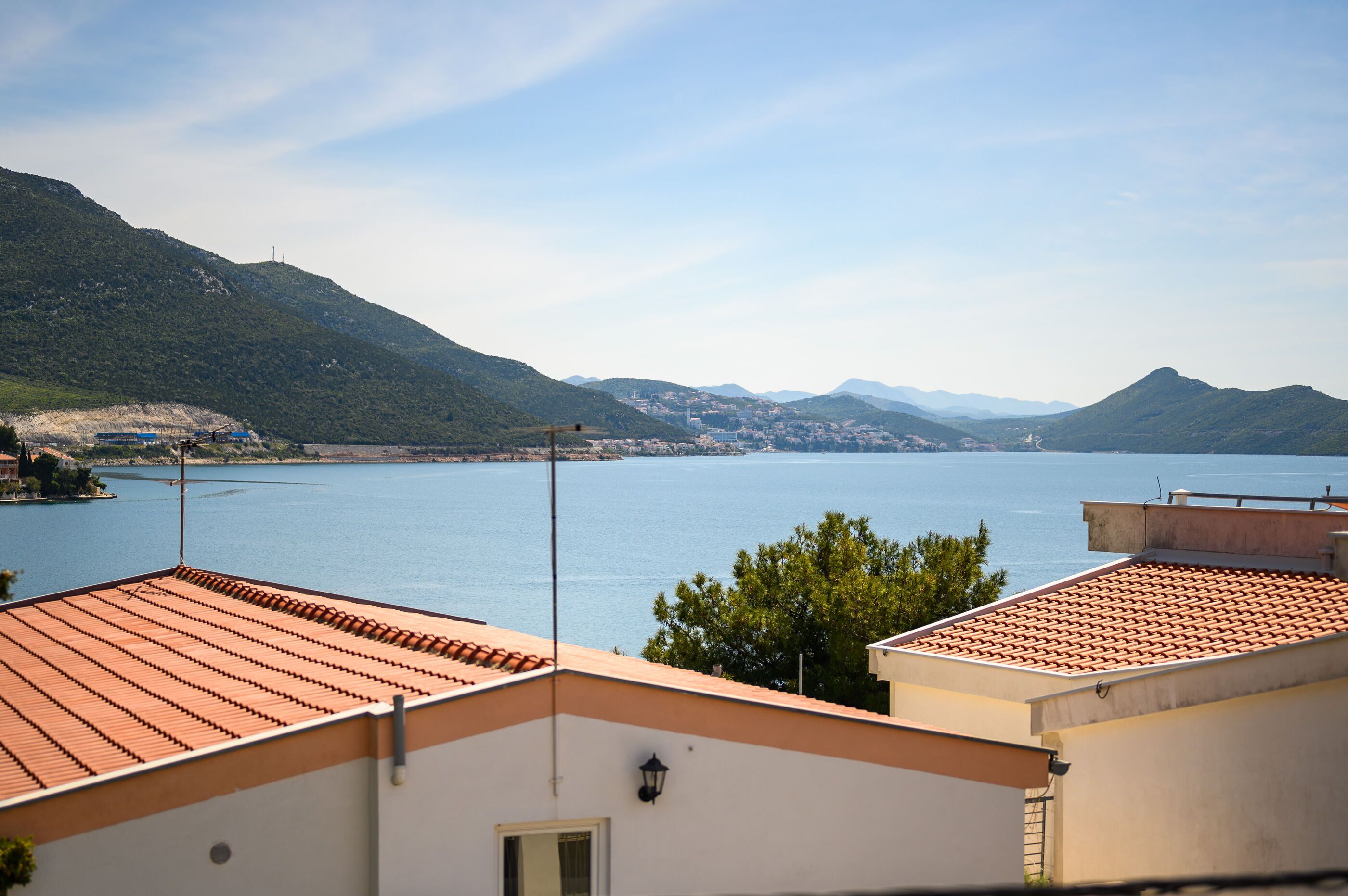 A coastal view with terracotta-roofed buildings overlooking a calm sea and distant mountains.