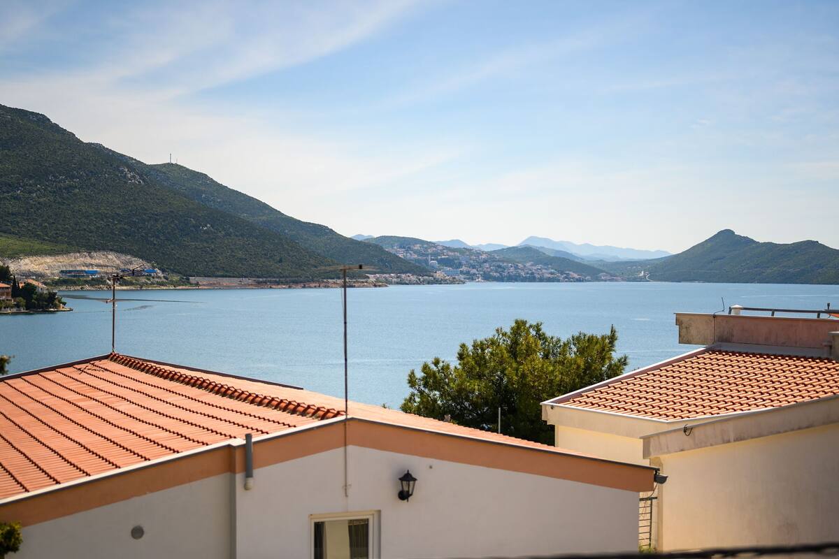 A coastal view with terracotta-roofed buildings overlooking a calm sea and distant mountains.