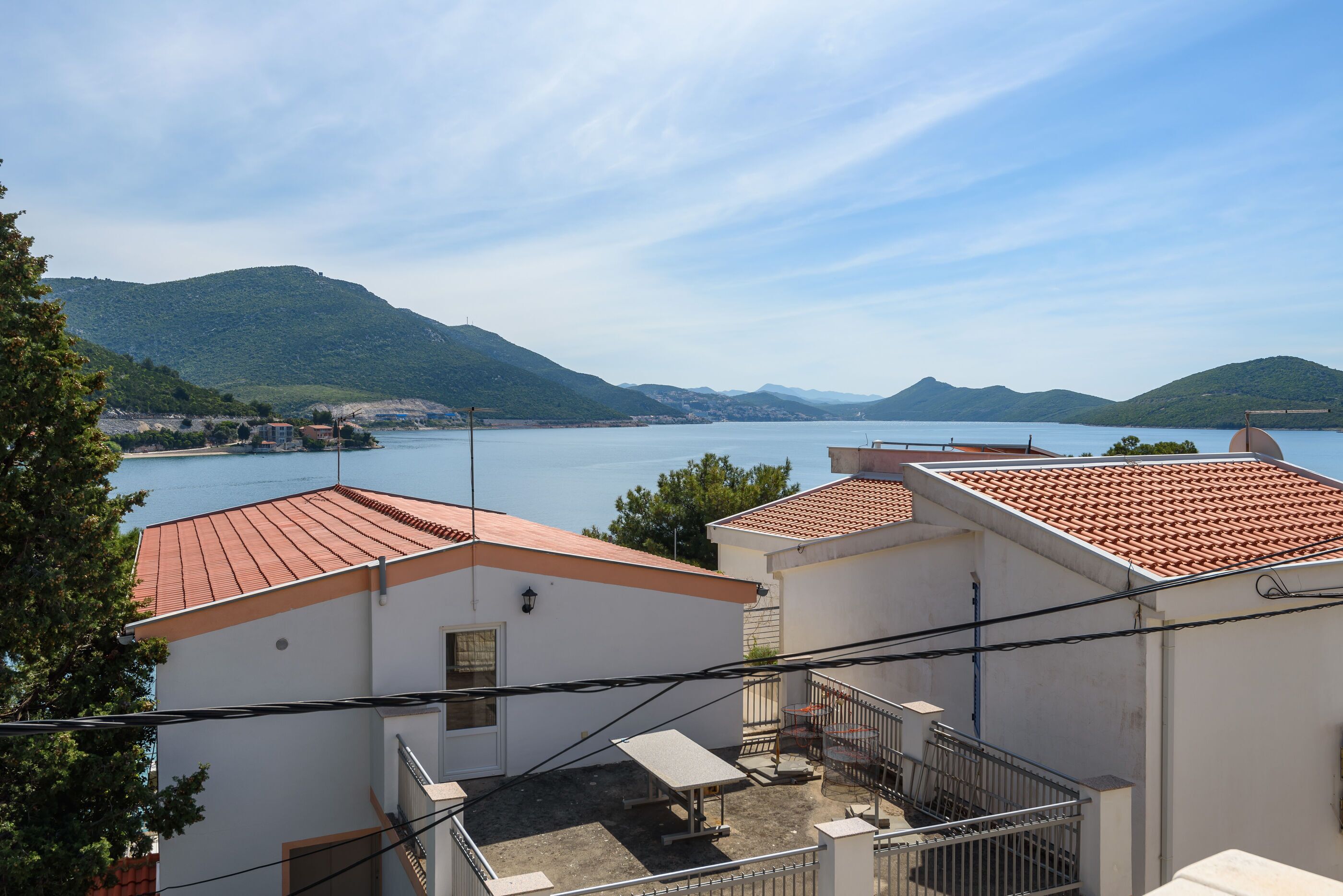 A coastal building with a red-tiled roof, a small balcony, and a view of the sea and hills in the background.