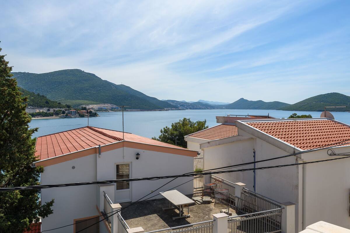 A coastal building with a red-tiled roof, a small balcony, and a view of the sea and hills in the background.