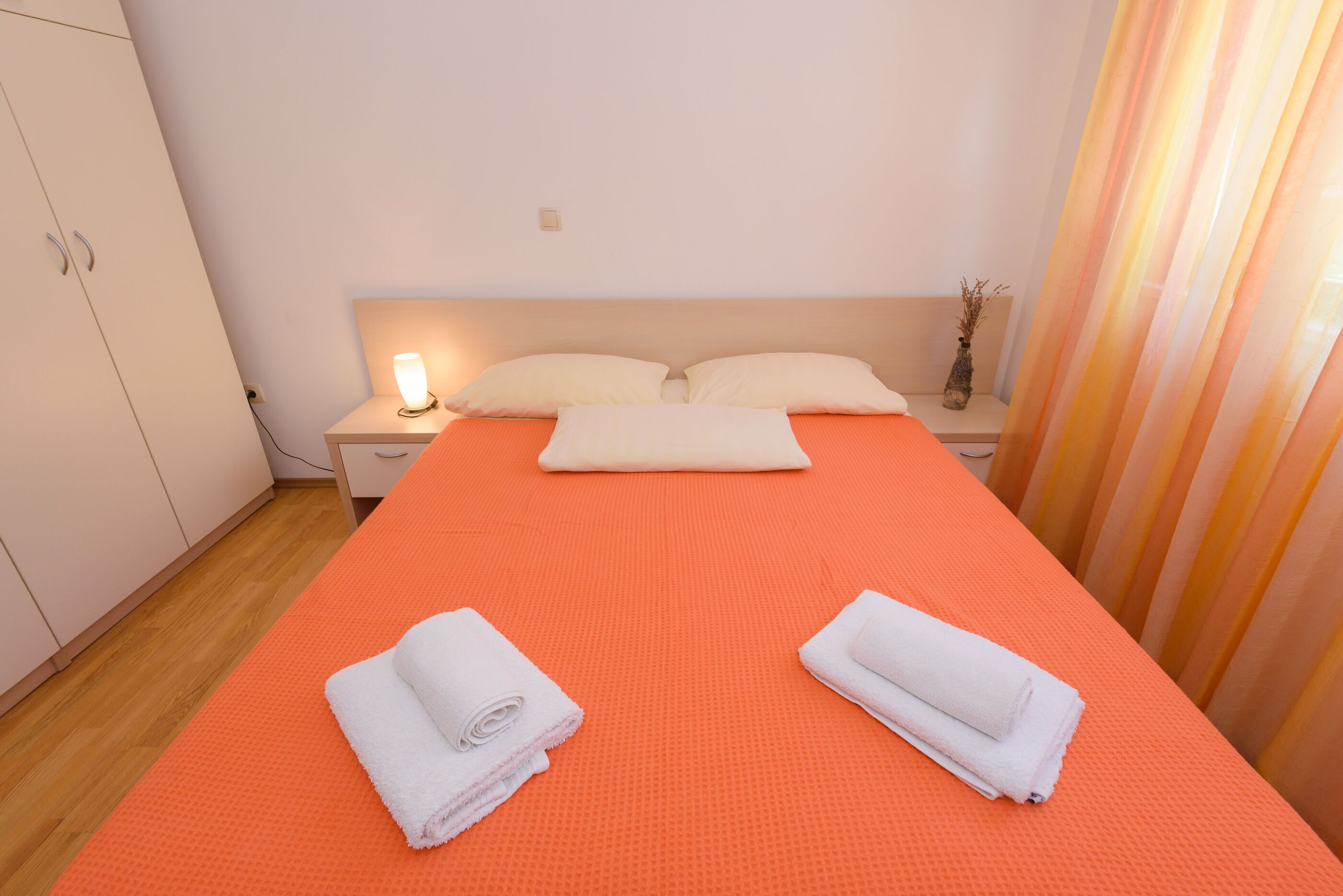Hotellihuone, jossa on yksi sänky, kaksi tyynyä, yöpöytä lampun kanssa ja yökaappi.