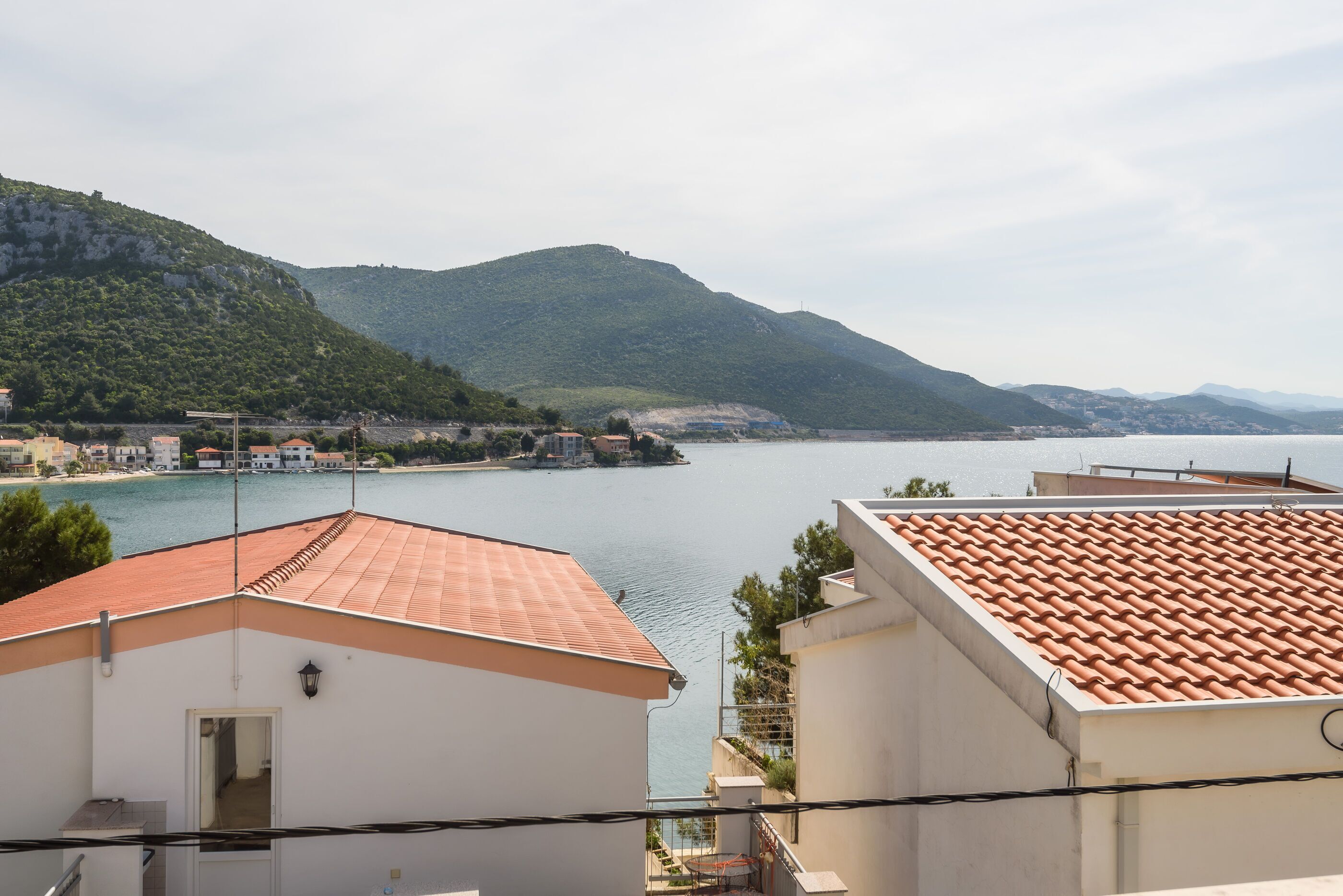 A coastal town with terracotta-roofed buildings, a clear view of the sea, and green hills in the background.