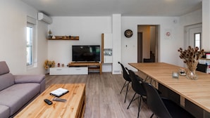 A modern living room with a sofa, a coffee table, a TV, a dining table with chairs, and a kitchen area with a fridge and a clock on the wall. - House Villa Ivona (Suhovare)
