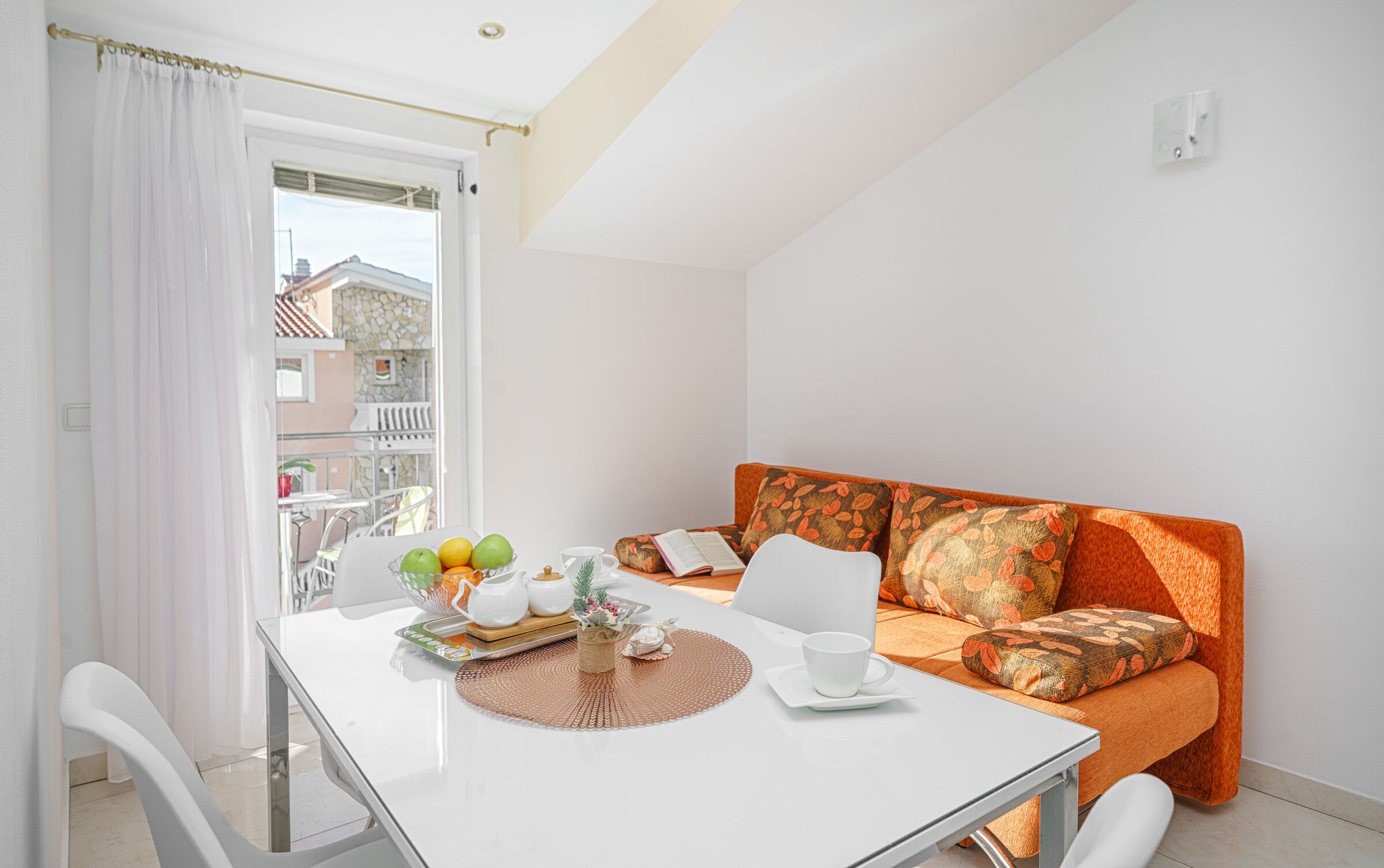 A bright room with a white table, a sofa with orange cushions, a window with white curtains, and a view of buildings outside.
