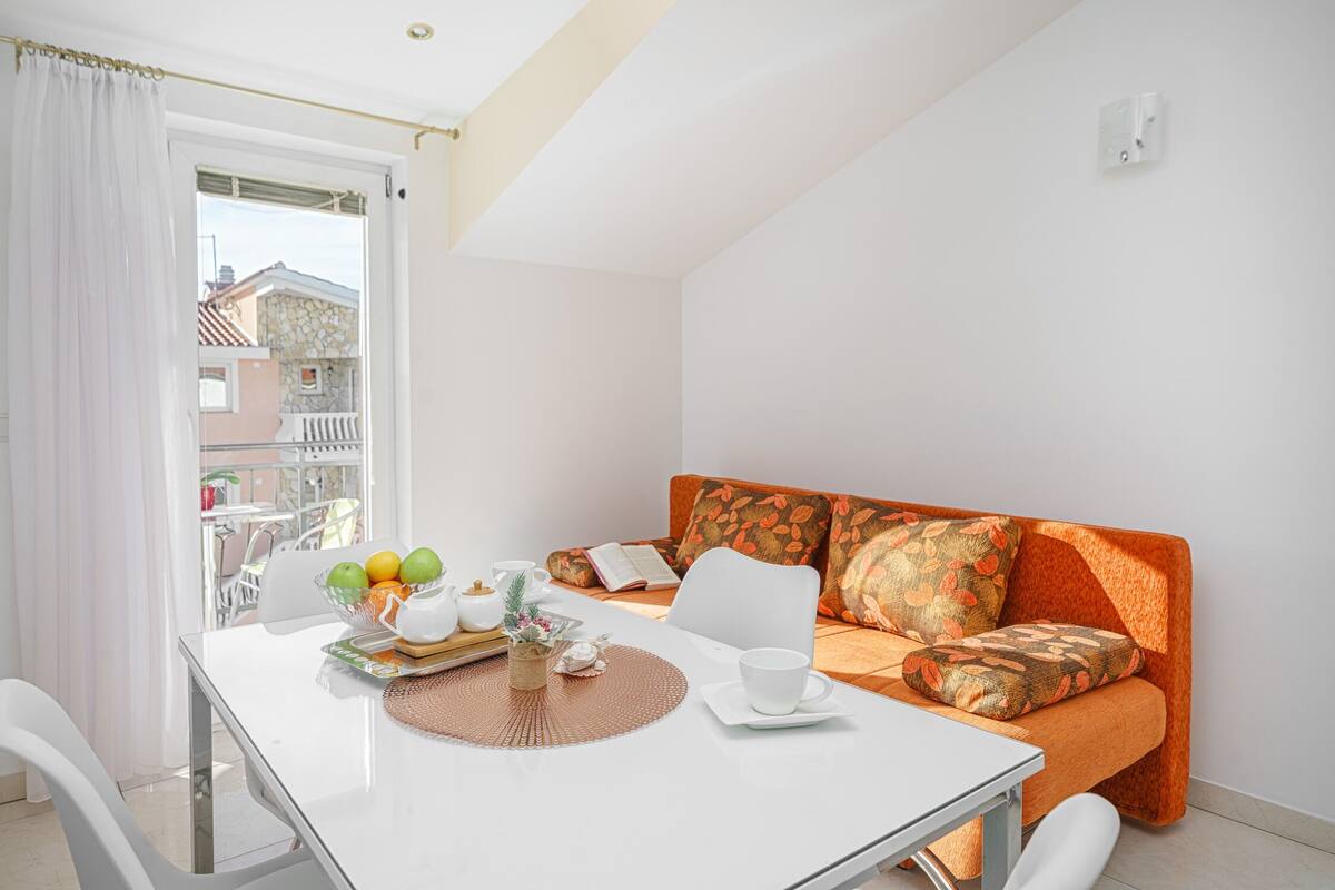 A bright room with a white table, a sofa with orange cushions, a window with white curtains, and a view of buildings outside.