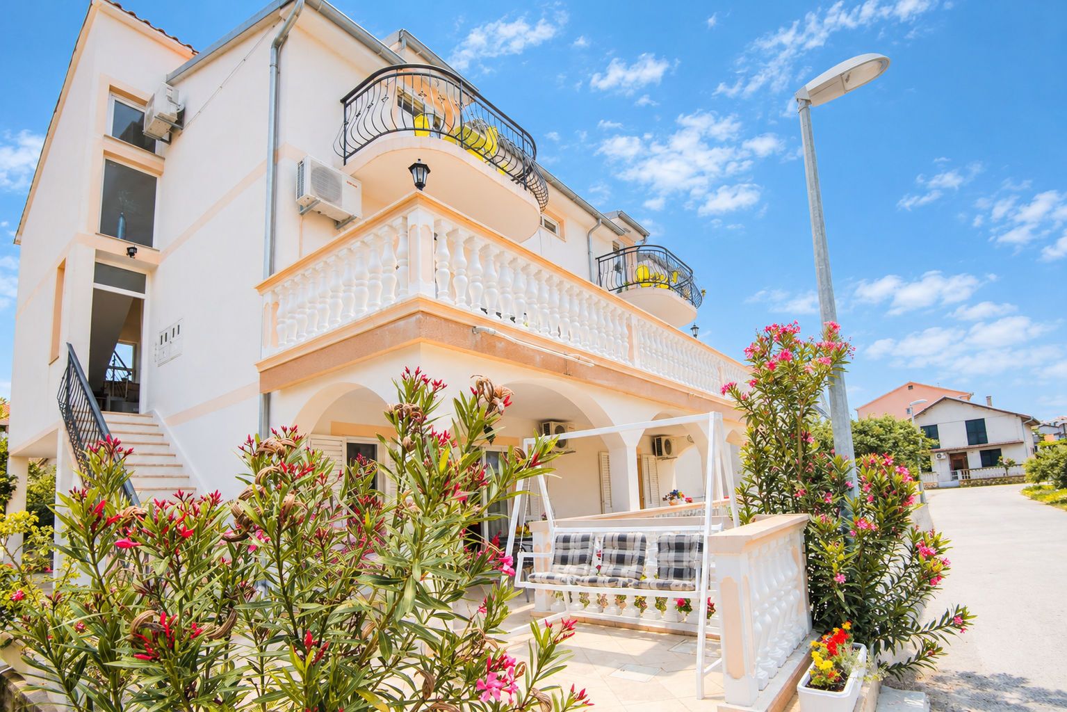 A two-story building with a balcony, a staircase, and a garden with flowering plants.