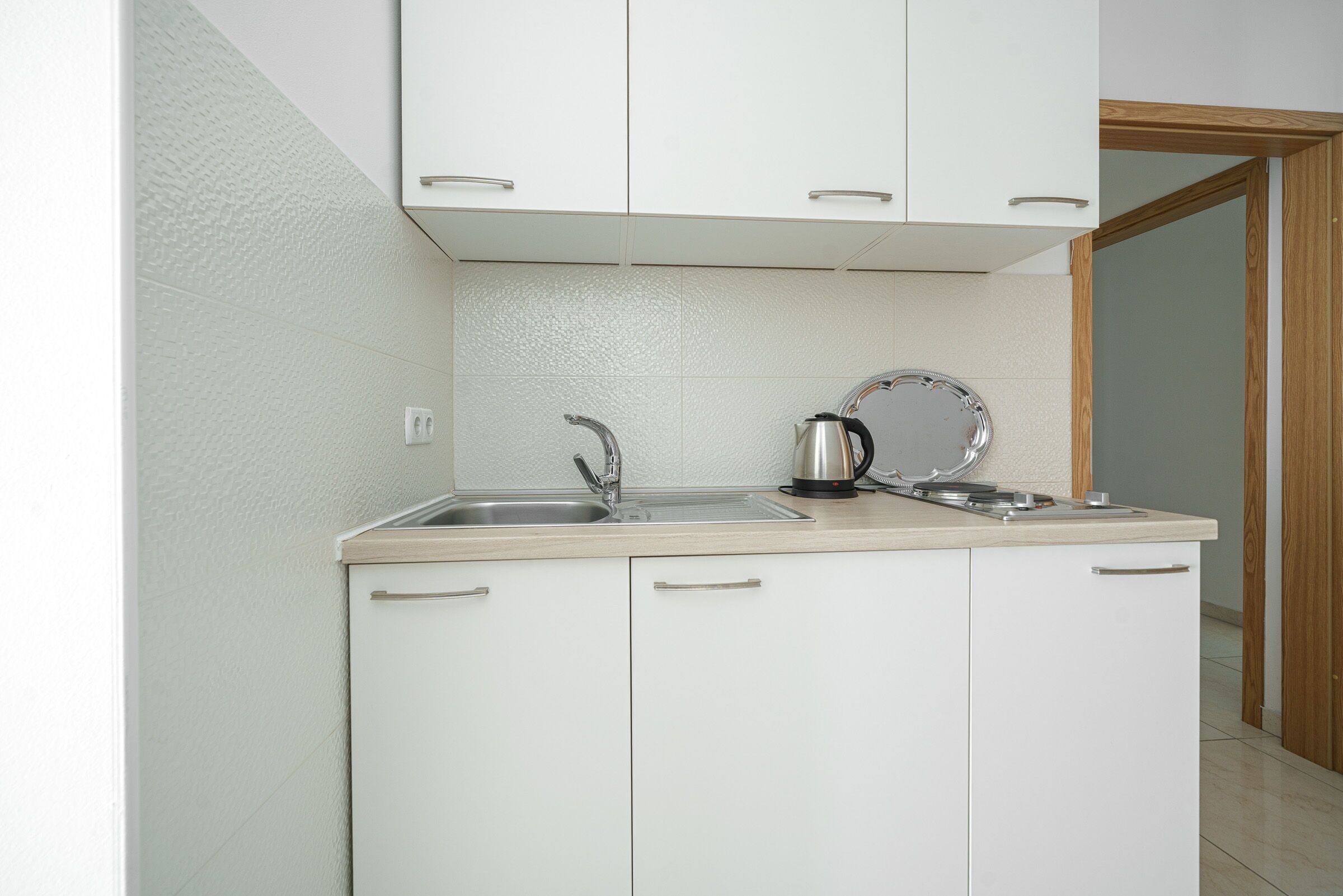 Una pequeña cocina con armarios blancos, un lavamanos y una tetera sobre la encimera.