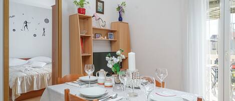 Un coin repas bien aménagé, avec une table dressée pour quatre personnes, un buffet en bois agrémenté d’objets décoratifs, et un lit avec une couette blanche en arrière-plan.
