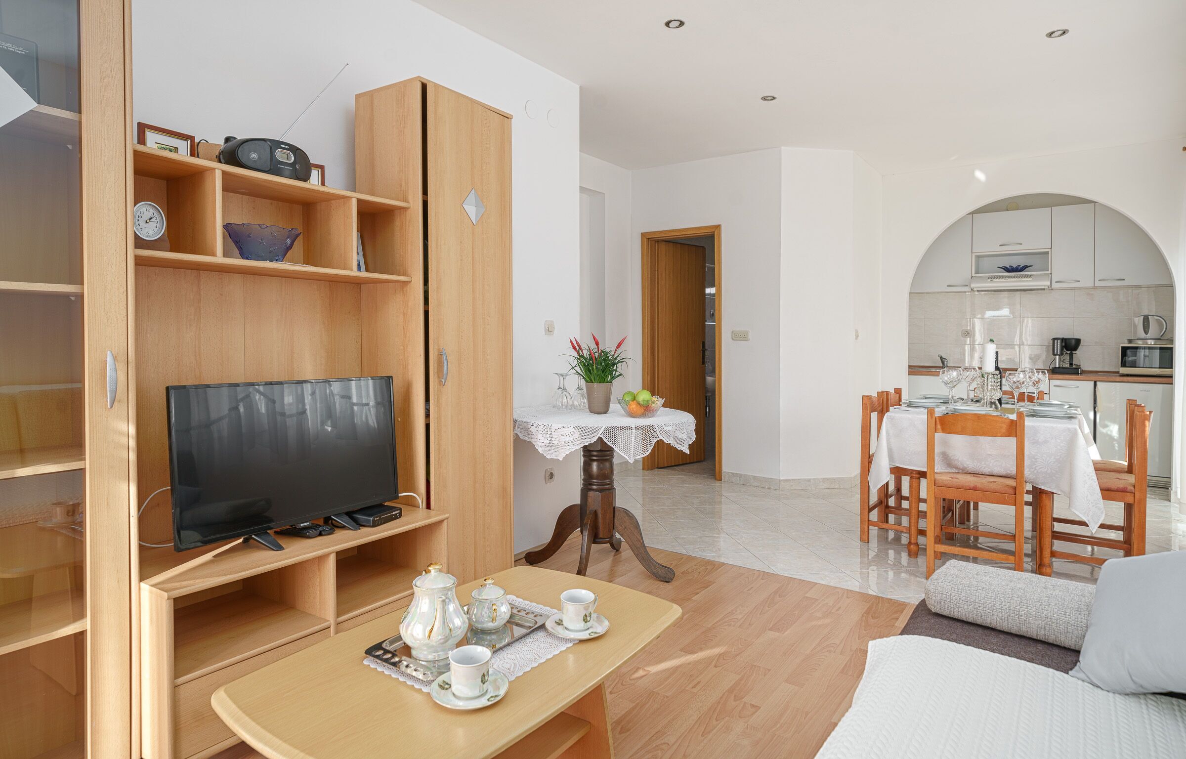 A modern living room with a TV, wooden shelves, a dining area with a table and chairs, and a kitchen in the background.