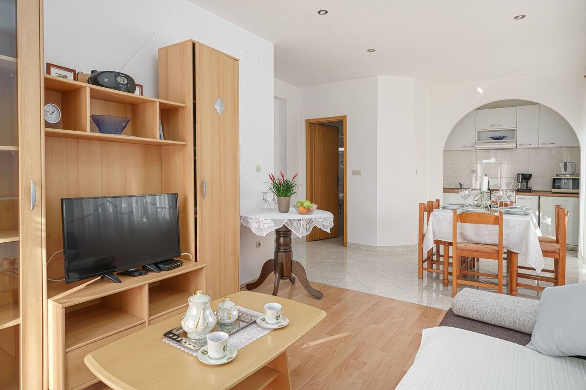 A modern living room with a TV, wooden shelves, a dining area with a table and chairs, and a kitchen in the background.