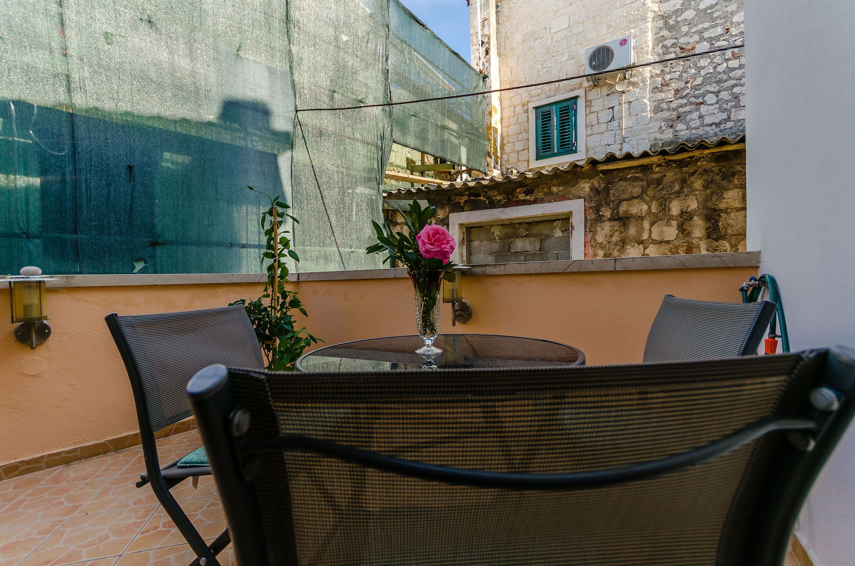 A patio with a round table, two chairs, a vase with a flower, and a wall-mounted air conditioning unit.