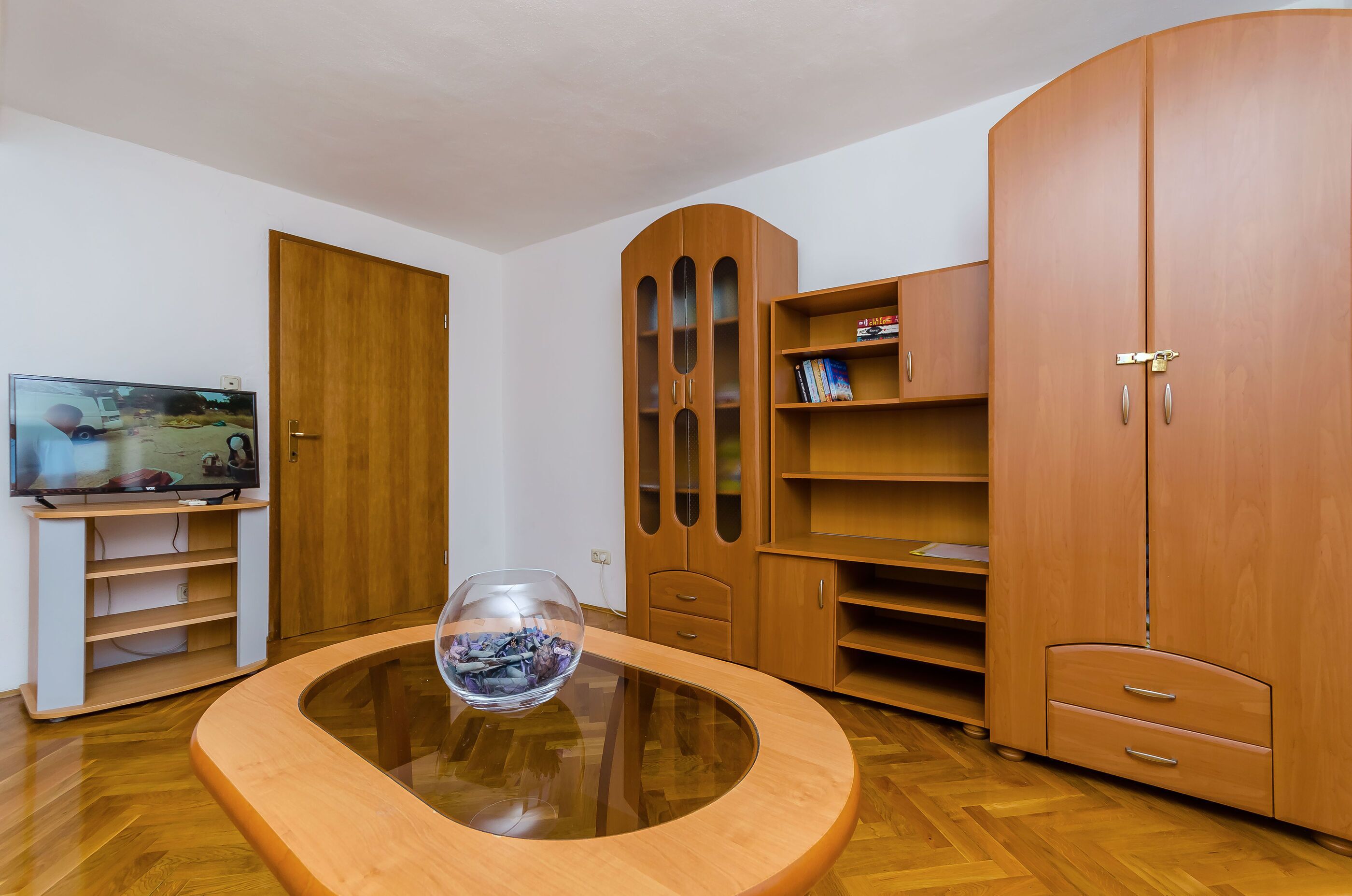 A modern living room with a glass-top coffee table, wooden cabinets, a television, and a door.