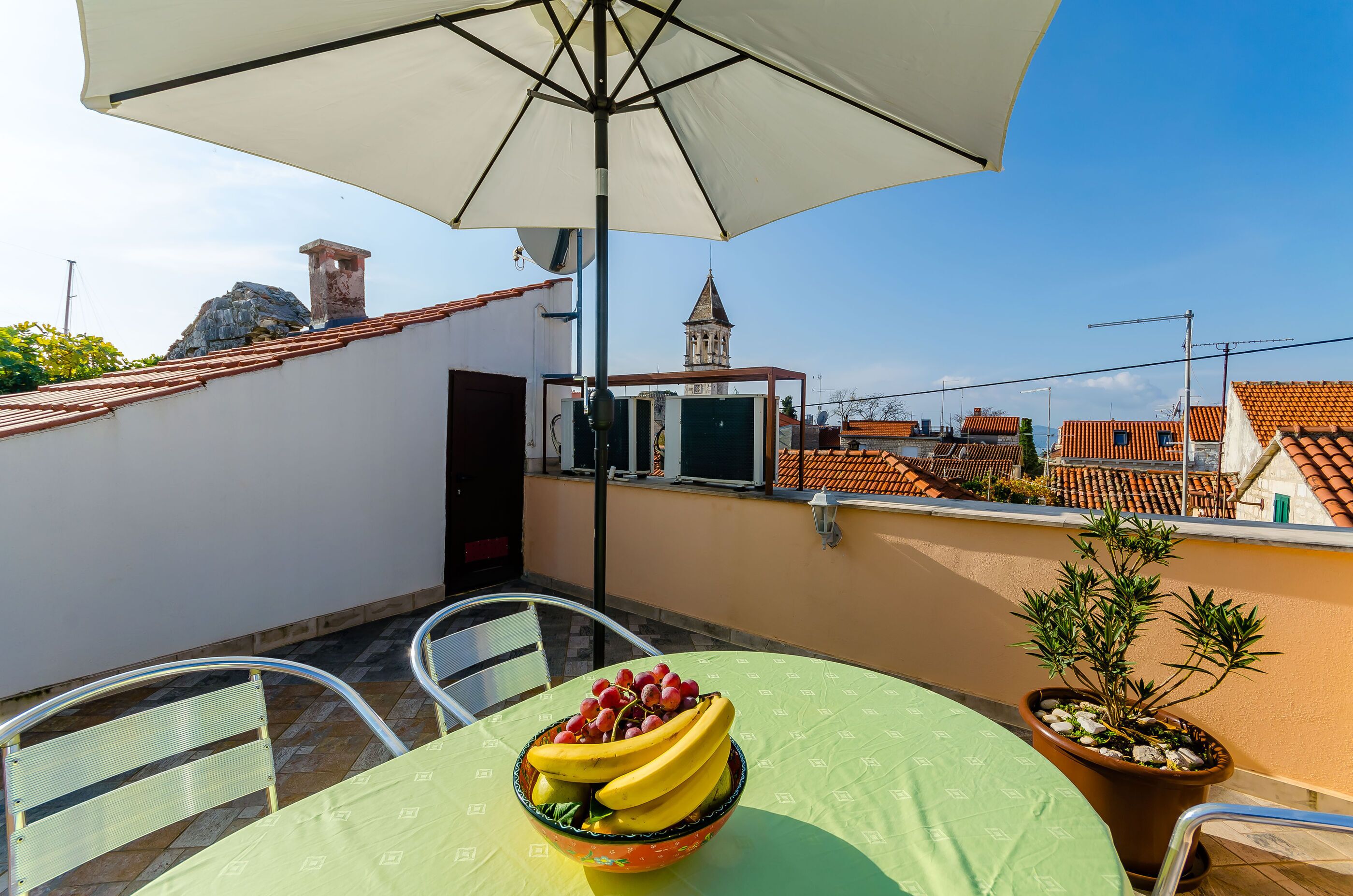 A balcony with a table, chairs, a bowl of fruit, a potted plant, and a view of buildings and a tower.