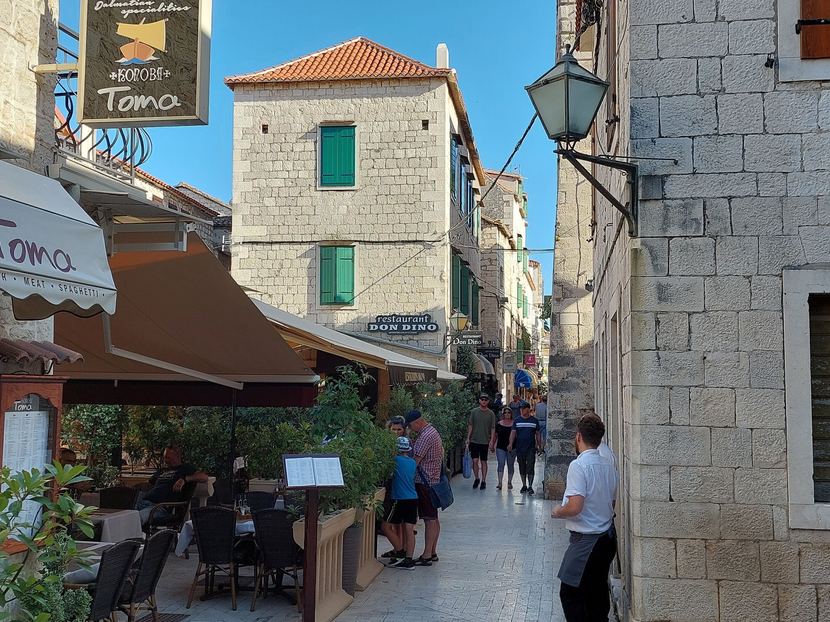 A narrow street with outdoor seating at a restaurant, a stone building with a tiled roof, and a sign for 'Don Dino' restaurant.