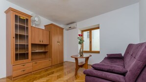 A living room with a wooden cabinet, a purple sofa, a small wooden table with a vase, and a window with blinds. - Apartments Old Town (Trogir)
