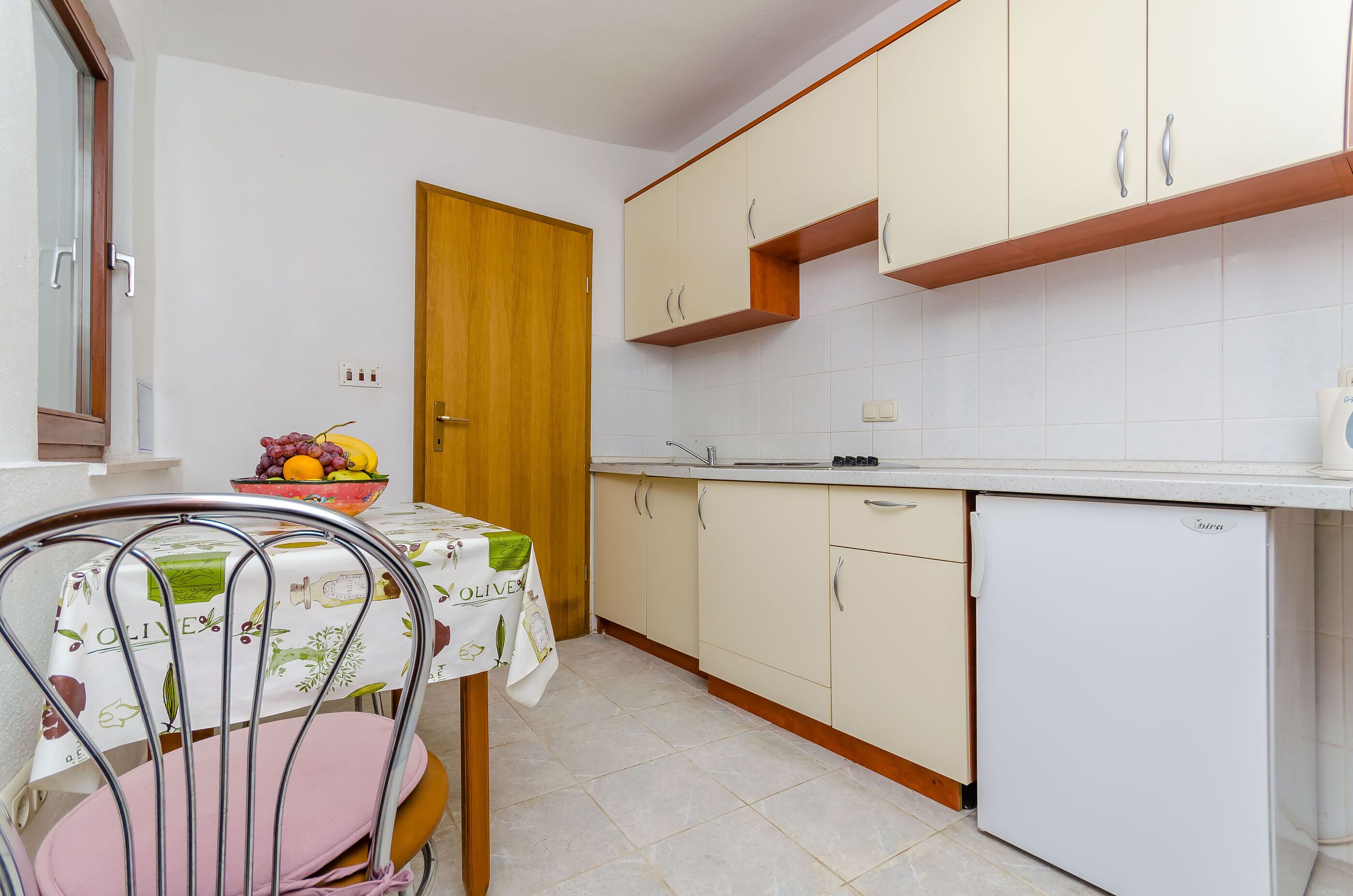 A compact kitchen with a dining table, a fruit bowl, and a refrigerator.