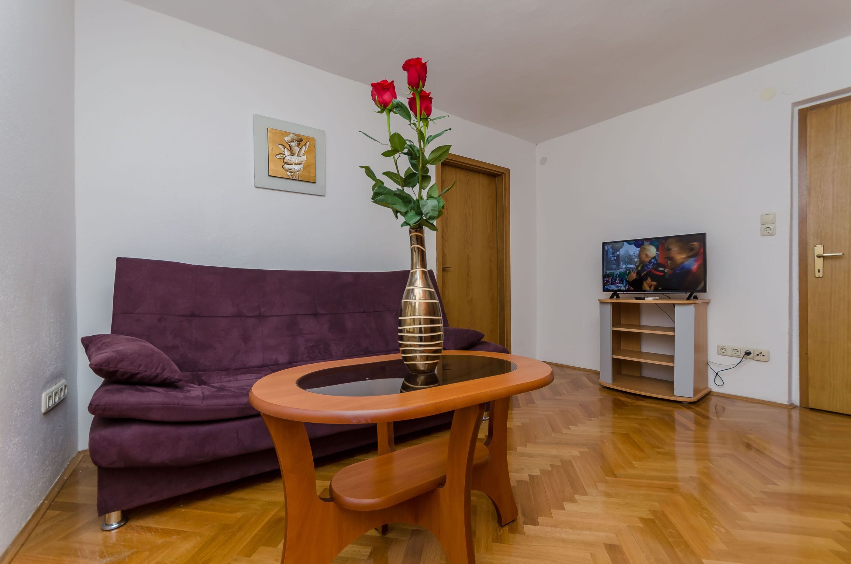 Ein Wohnzimmer mit einem violetten Sofa, einem Holztisch mit einer Vase voller roter Rosen, einem Fernseher auf einem Schrank und einer Holztür.