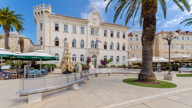 A public square with a large building, outdoor seating, and palm trees. - Apartments Old Town (Trogir)