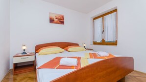 A bedroom with a wooden bed, a nightstand with a lamp, a window with curtains, and a painting on the wall. - Apartments Old Town (Trogir)