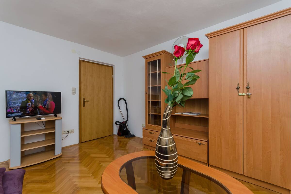 A living room with a TV, wooden cabinets, a glass coffee table, and a vase with a rose.