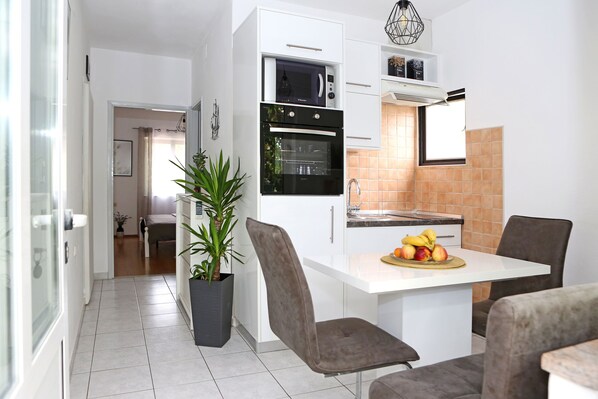 Una cocina moderna con armarios blancos, horno integrado y microondas. Hay una zona de comedor con mesa y sillas, y una planta en maceta.