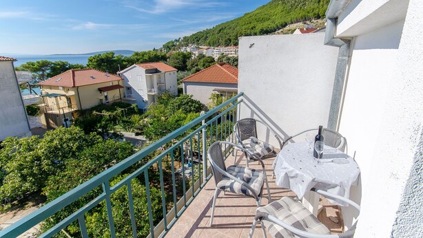 A balcony with wicker chairs and a table, overlooking a hillside with houses and a body of water.