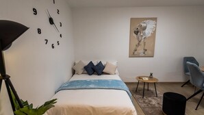 A bedroom with a bed, a clock, a painting, a chair, a small table, and a plant. - Apartment N D (Sibenik)