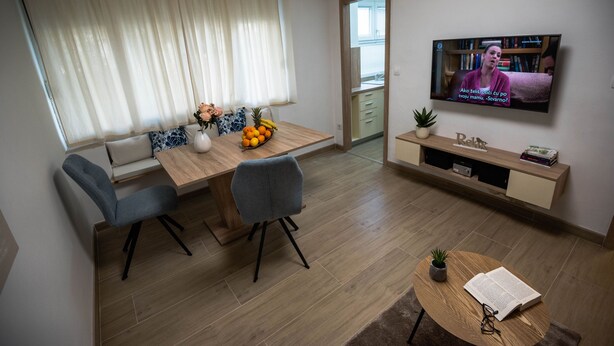 A modern living room with a wooden dining table, two grey chairs, a TV mounted on the wall, and a small round table with a book and glasses. - Apartment N D (Sibenik)