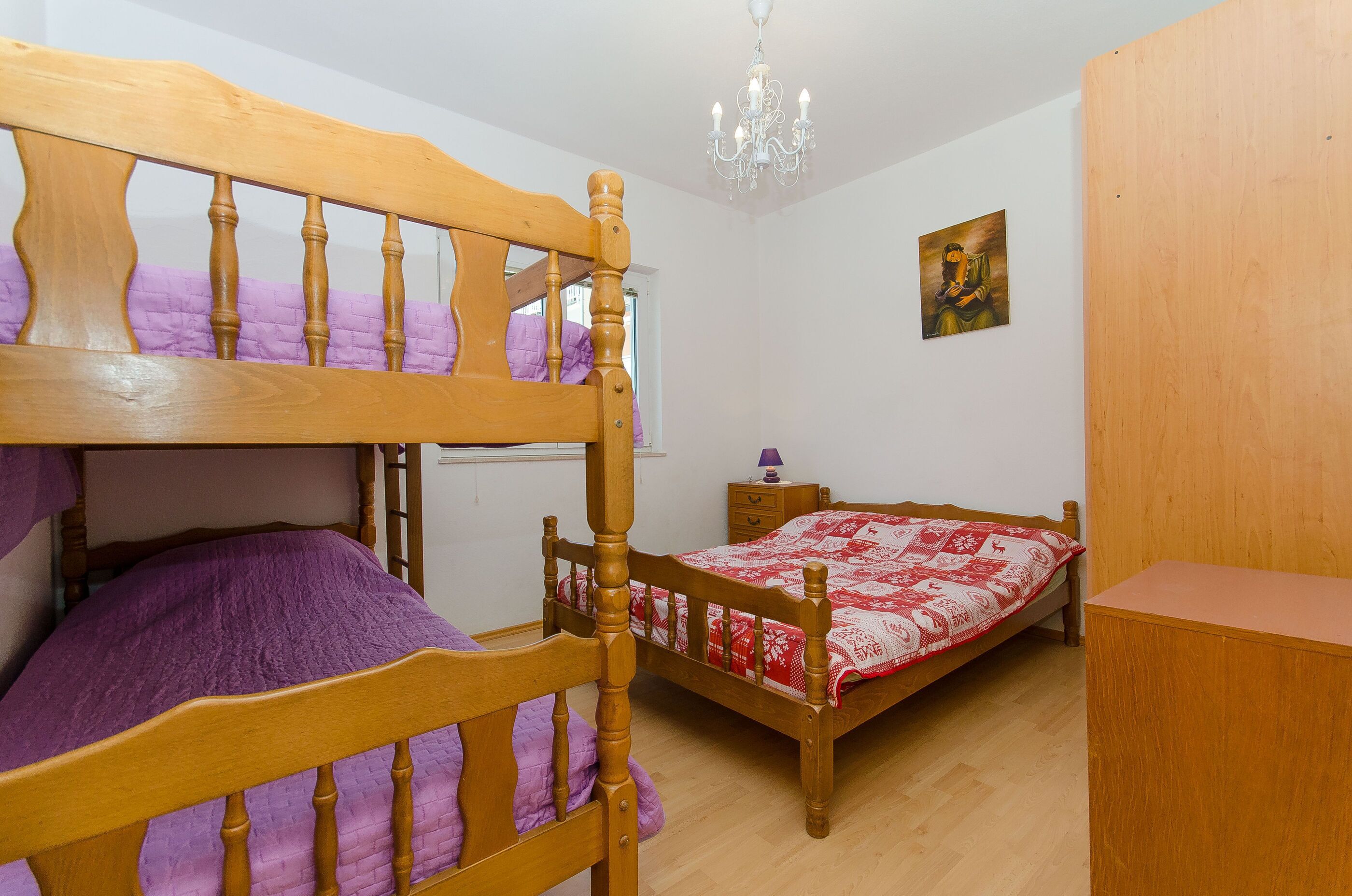 A bedroom with two wooden bunk beds, a wooden desk, a nightstand, a chandelier, and a wall painting.