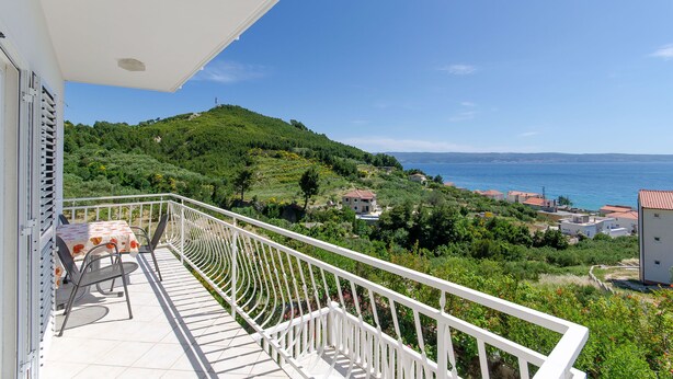 A balcony with a table and chairs, overlooking a coastal landscape with houses and greenery. - Apartment Marija (Podstrana)