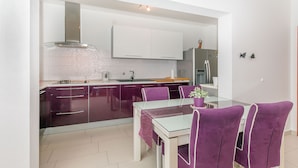 A modern kitchen with a glass dining table and purple chairs, white cabinets, and a stainless steel refrigerator. - Apartment Mario (Strozanac)