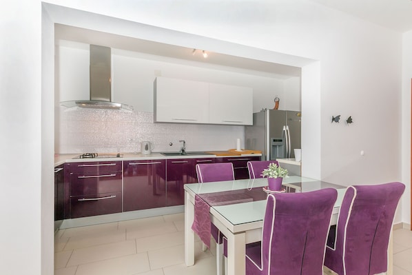 A modern kitchen with dark cabinets, a white countertop, and a glass dining table with purple chairs.