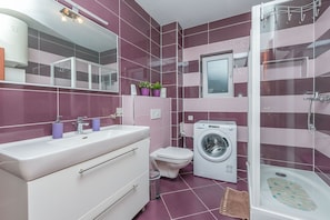 A modern bathroom with a large mirror, a sink, a toilet, a washing machine, and a shower area. - Apartment Mario (Strozanac)